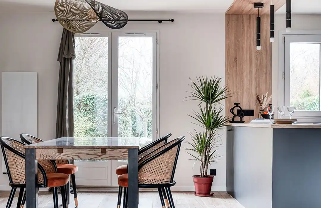 Rénovation séjour Un coin repas moderne, conçu dans le cadre d'un séjour de rénovation récente, comprend une table en verre et quatre chaises tressées. De grandes fenêtres inondent l'espace de lumière, tandis qu'une plante en pot et des lampes suspendues lui confèrent un style élégant.